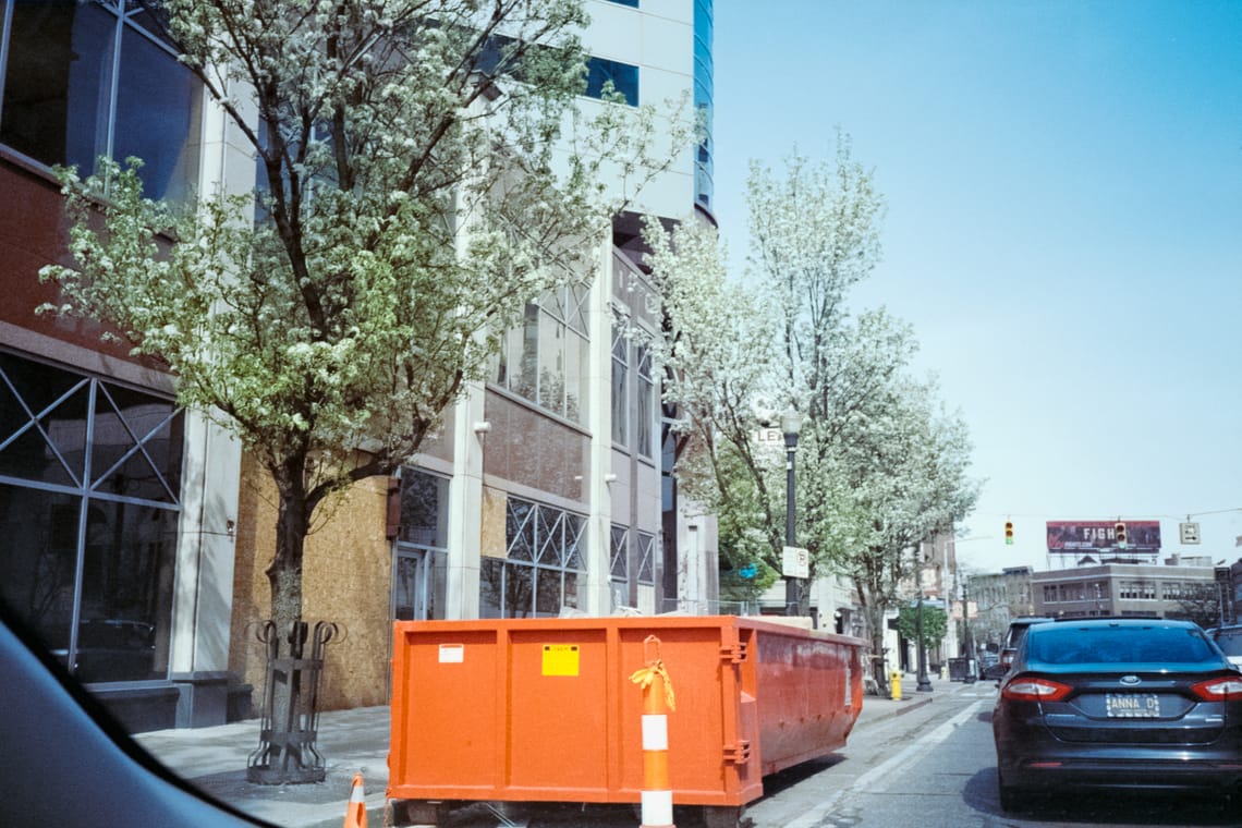 A shot from within a car of a flowering row of trees along a road with a large orange dumpster in the foreground.