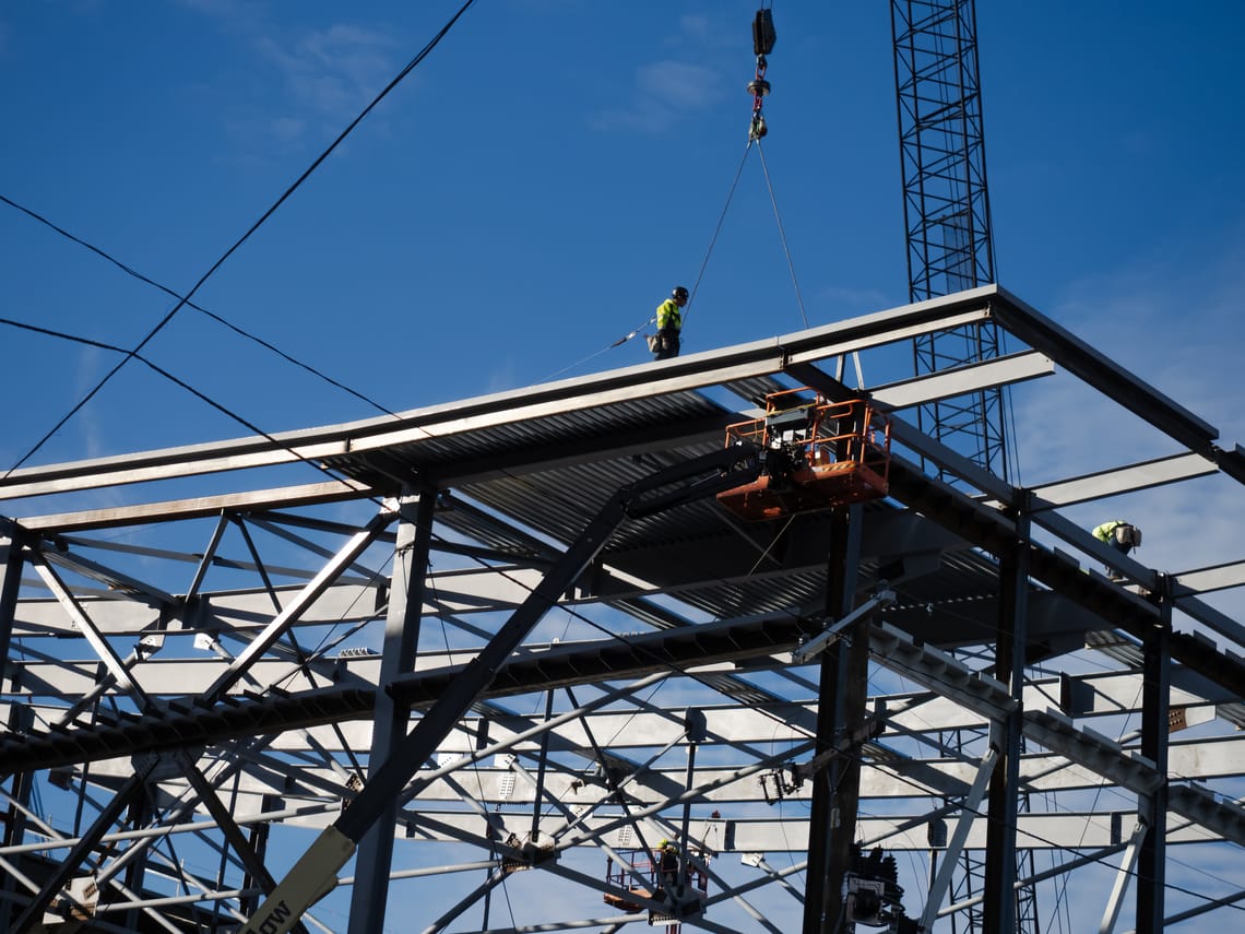 Construction workers on top of the angular, metal frame of a building with a crane in the background.