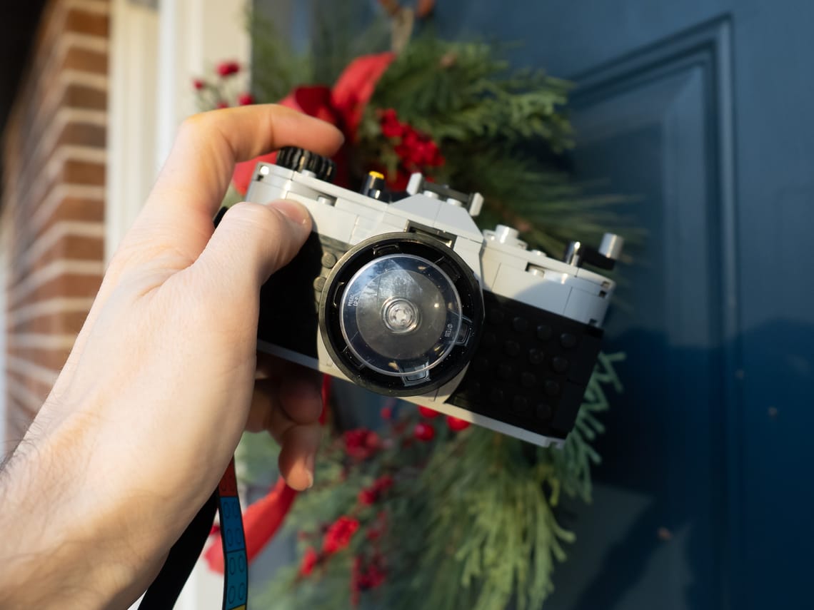 A lego camera with a reflection of a pine tree in the "lens" and a holiday wreath on a door in the background.