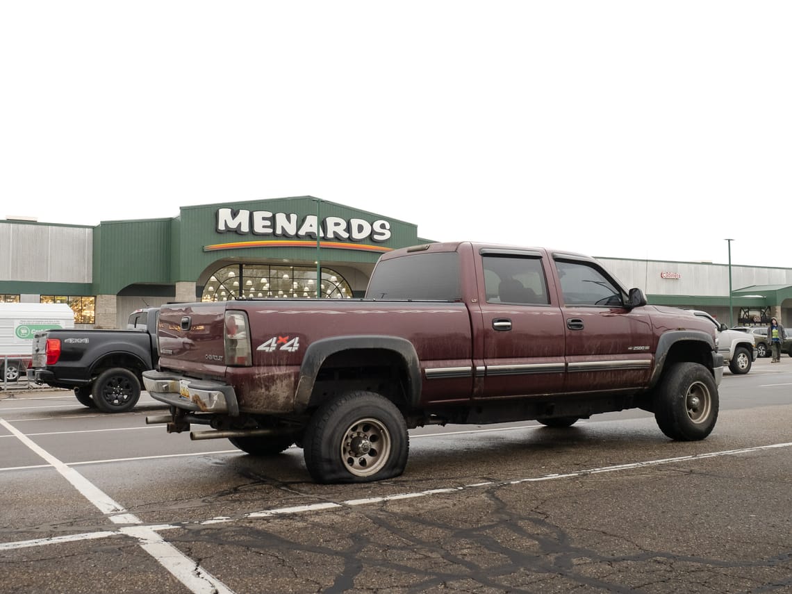 A maroon 4x4 pickup truck in a Menards parking lot with a flat tire. A man is walking towards the camera in the distance (right).