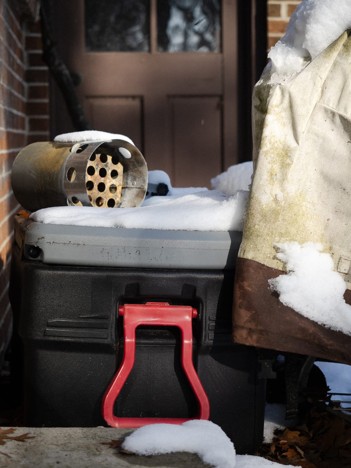 A red bin with snow on it. The sun is lighting the side. There is a charcoal grill chimney starter on top the bin. A cloth is draped on the right side of the frame.