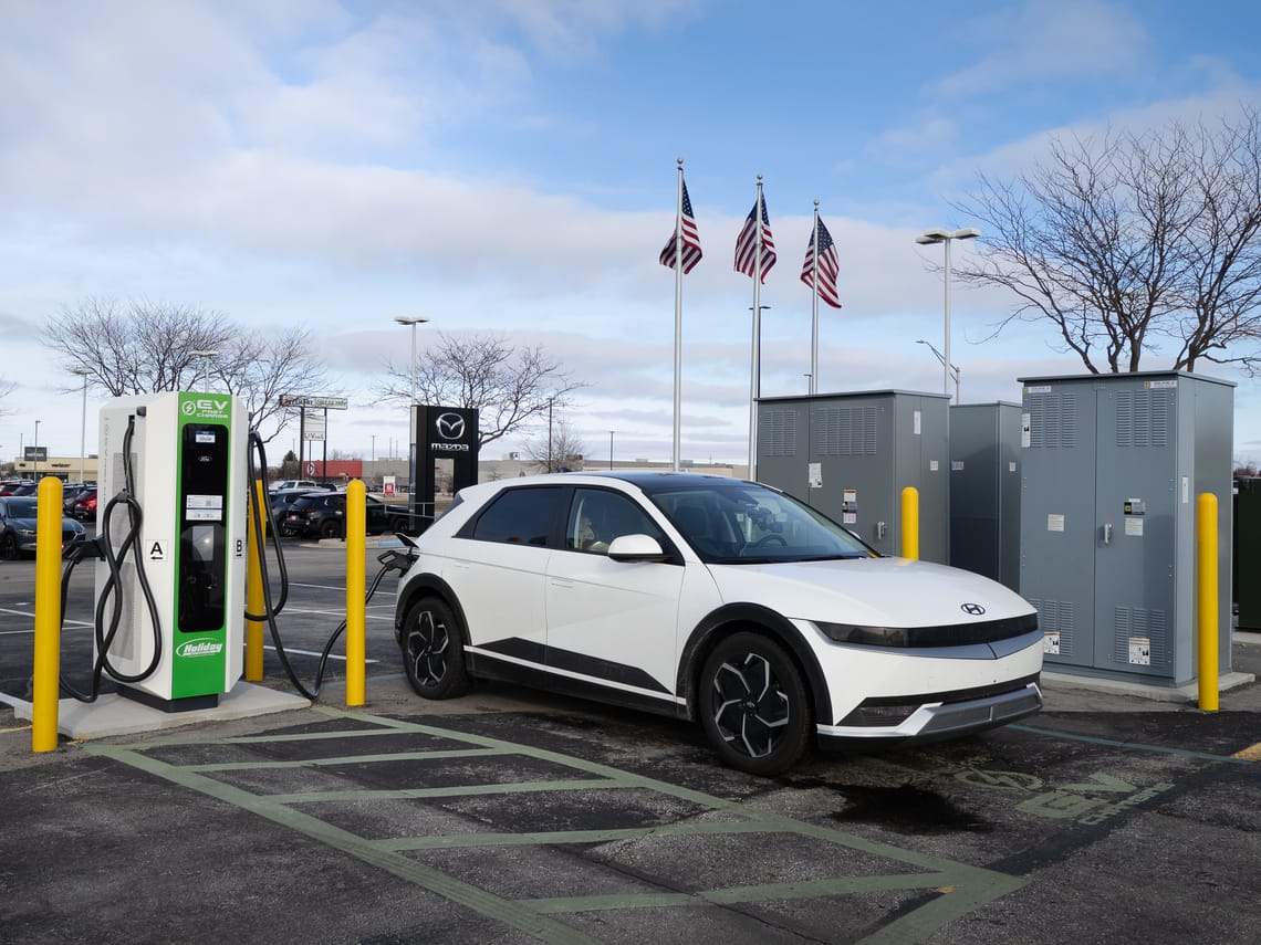 A white Hyundai IONIQ 5 EV charges in front of a Mazda dealership sign. There are yellow bollards protecting the charger infrastructure.