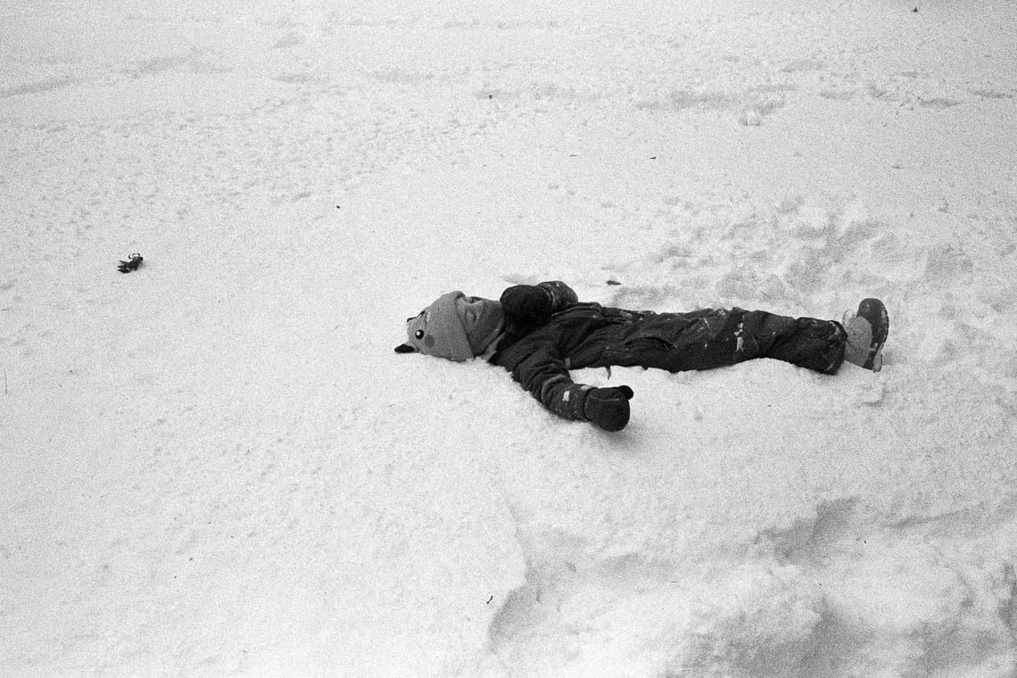 A child in a snow suit wearing a pikachu beanie lies in the snow. The photo is a black and white film photo with lots of visible grain.