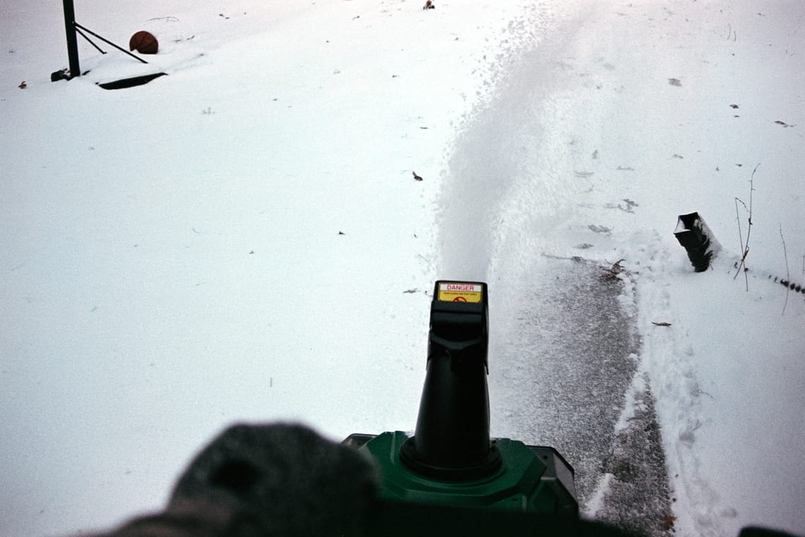 A POV perspective of a green and black snow blower with a gloved hand in the bottom left of the frame.