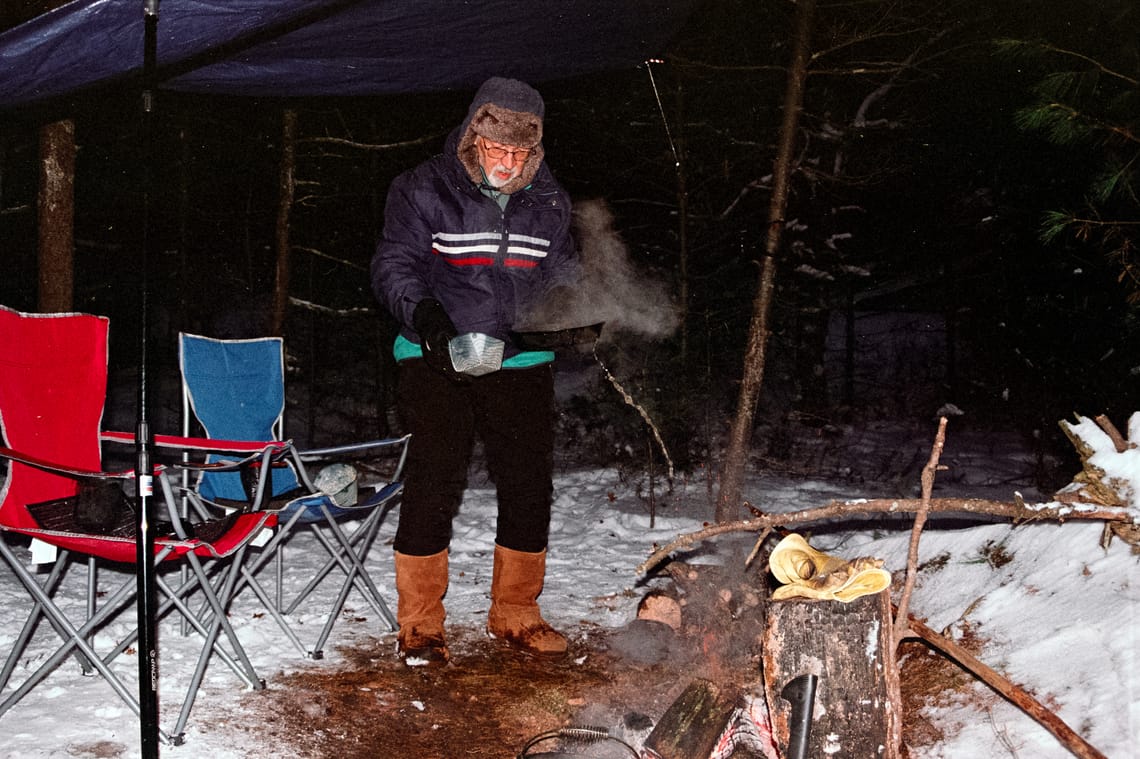 A man at a camp site with a fire tosses some liquid out of a bowl onto the ground. Two chairs are visible on the left of the frame.