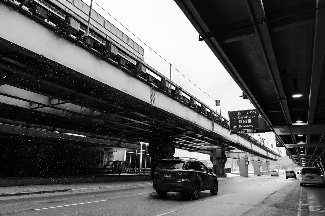 A black and white photo of the pick-up area of Chicago O'Hare airport. Snow falling is visible as cars drive by.