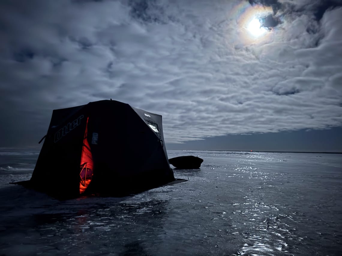 An insulated ice fishing tent anchored into the ice. The moon is illuminating the ice behind the tent.