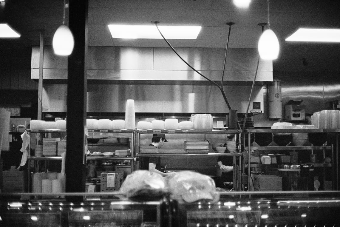 A black and white film photo of a restaurant kitchen with two works in the background behind some shelves. There are dangling light fixtures that add a cozy atmosphere to the scene.